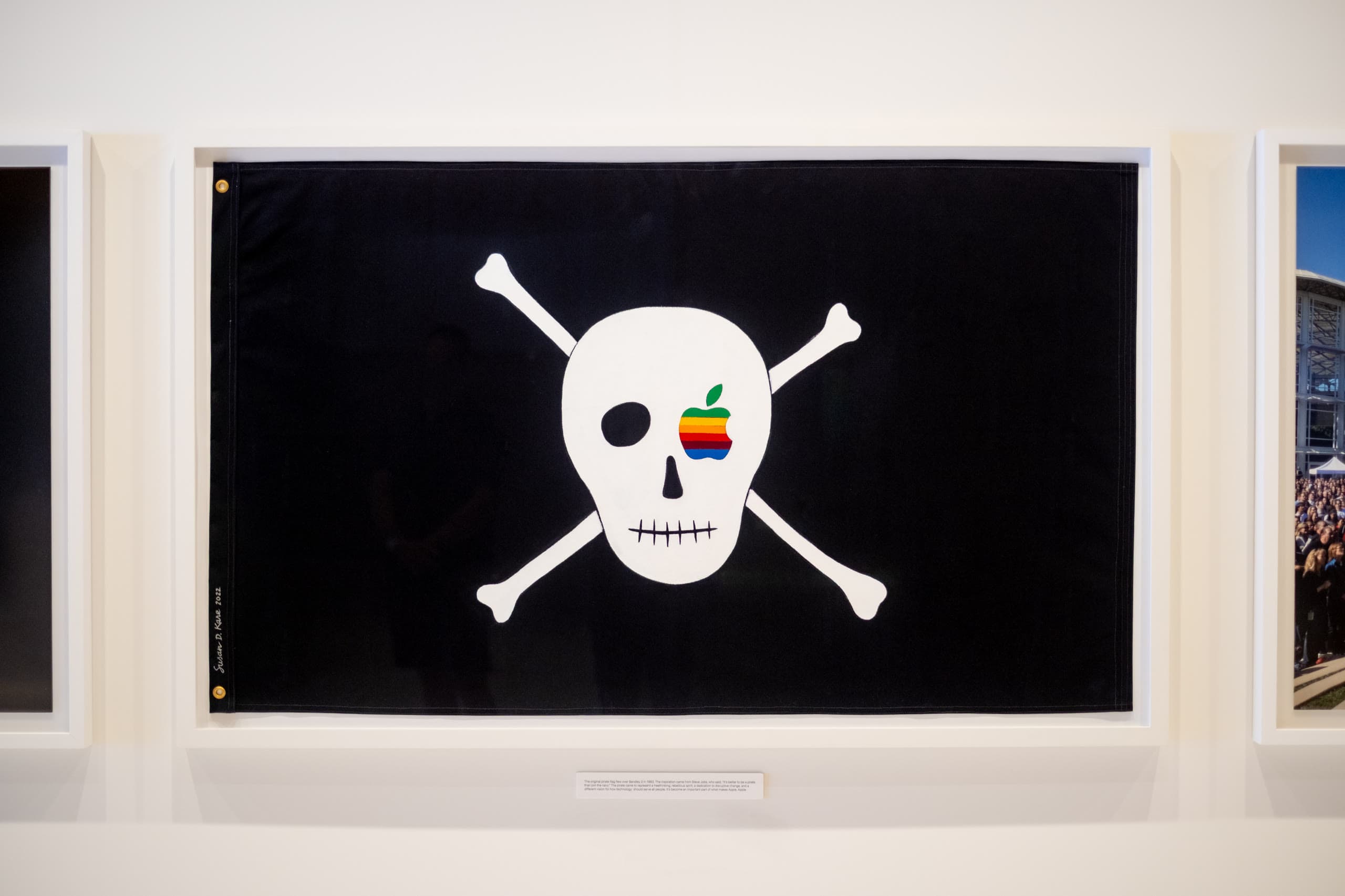 A framed black pirate flag with a white skull and crossbones, the eye replaced by the rainbow Apple logo, signed by Susan Kare
